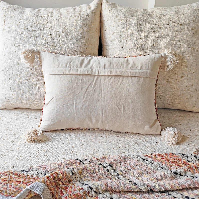 May include: A white throw pillow with tassels on the corners. The pillow is on a bed with a white textured bedspread and a multi-coloured throw blanket.