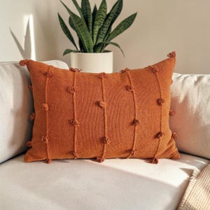 May include: A rectangular burnt orange throw pillow featuring a textured surface with vertical lines and small, evenly spaced tufted knots. The pillow has a woven fabric construction with a consistent solid color throughout the entire piece.