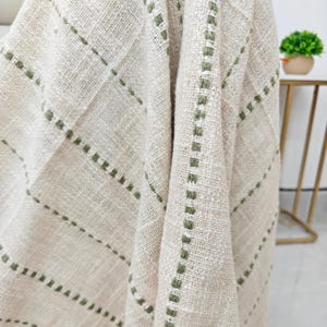 Puede incluir: Primer plano de una manta tejida beige con aspecto texturizado. La manta presenta un patrón de cuadrícula con costuras verde oliva. La tela está drapeada, mostrando la textura y el diseño. Una pequeña planta en maceta es visible en el fondo.