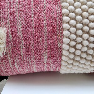 May include: A pink and white striped throw pillow with a textured surface. The pillow has a white pom-pom trim on one side.