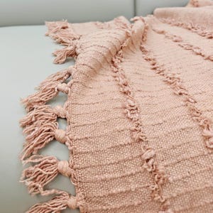May include: A close-up of a blush-colored woven throw blanket with a textured design. The blanket has knotted fringe along the edge. The woven pattern features vertical lines and a soft, neutral color.
