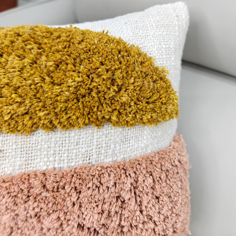 May include: Close-up of a decorative pillow with a textured design. The pillow features a mustard yellow, white, and dusty rose color scheme. The yellow and rose sections have a shaggy, raised texture, while the white section is a woven fabric.