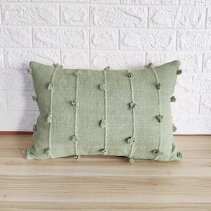 以下が含まれることがあります： 縁に小さなポンポンが付いた、緑がかった灰色の質感のある生地のクッションです。