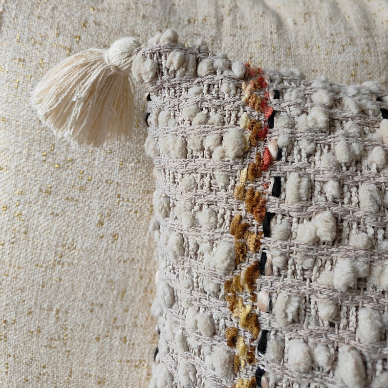 May include: A close-up of a cream-coloured throw pillow with a textured woven design. The pillow has a fringe trim and a small cream-coloured tassel.