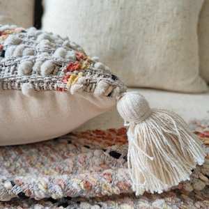 May include: A cream-coloured throw pillow with a textured woven design and a fringe tassel. The pillow has a subtle pattern of orange, yellow, and brown.