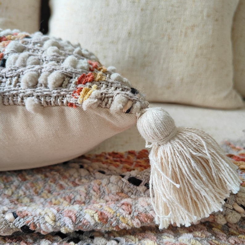 May include: A cream-coloured throw pillow with a textured woven design and a fringe tassel. The pillow has a subtle pattern of orange, yellow, and brown.