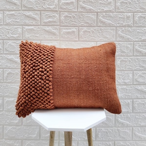 May include: A rust-colored throw pillow with a textured woven fabric and a fringe detail on one side. The pillow is sitting on a white hexagonal table.