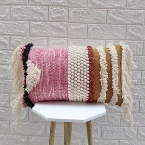 May include: A pink, white, and brown throw pillow with fringe detailing. The pillow has a textured woven design and is made of soft materials.