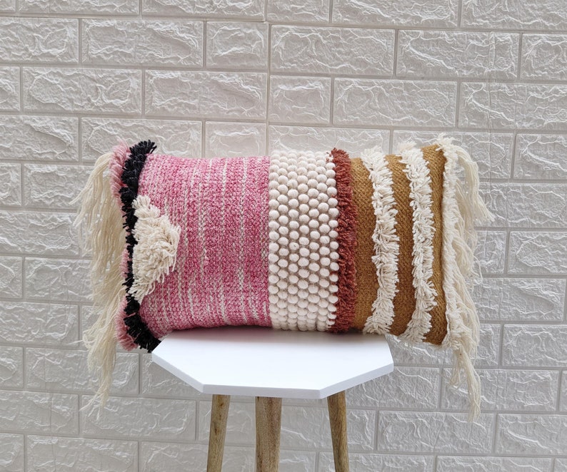 May include: A pink, white, and brown throw pillow with fringe detailing. The pillow has a textured woven design and is made of soft materials.