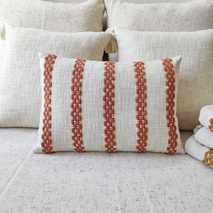 Puede incluir: Una almohada blanca con cuatro rayas naranjas tejidas en la tela. La almohada está sobre una colcha blanca texturizada.