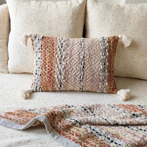 May include: A throw pillow with a woven pattern in shades of beige, orange, and brown. The pillow has tassels on each corner and is sitting on a white sofa.