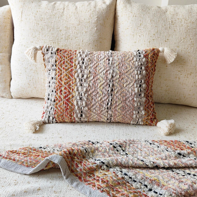 May include: A throw pillow with a woven pattern in shades of beige, orange, and brown. The pillow has tassels on each corner and is sitting on a white sofa.