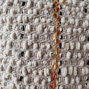 May include: Close-up of a beige woven fabric with a textured surface. The fabric has a vertical stripe of orange, yellow, and black thread.
