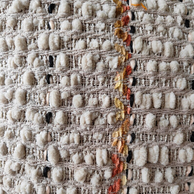 May include: Close-up of a beige woven fabric with a textured surface. The fabric has a vertical stripe of orange, yellow, and black thread.