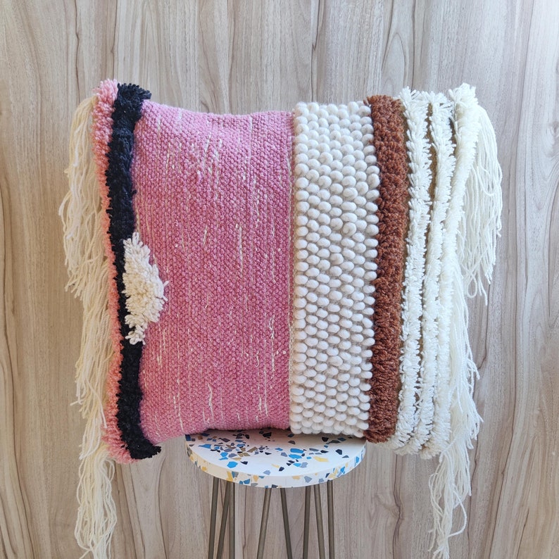 May include: A pink and white throw pillow with a fringe border. The pillow has a textured design and is made of woven fabric.