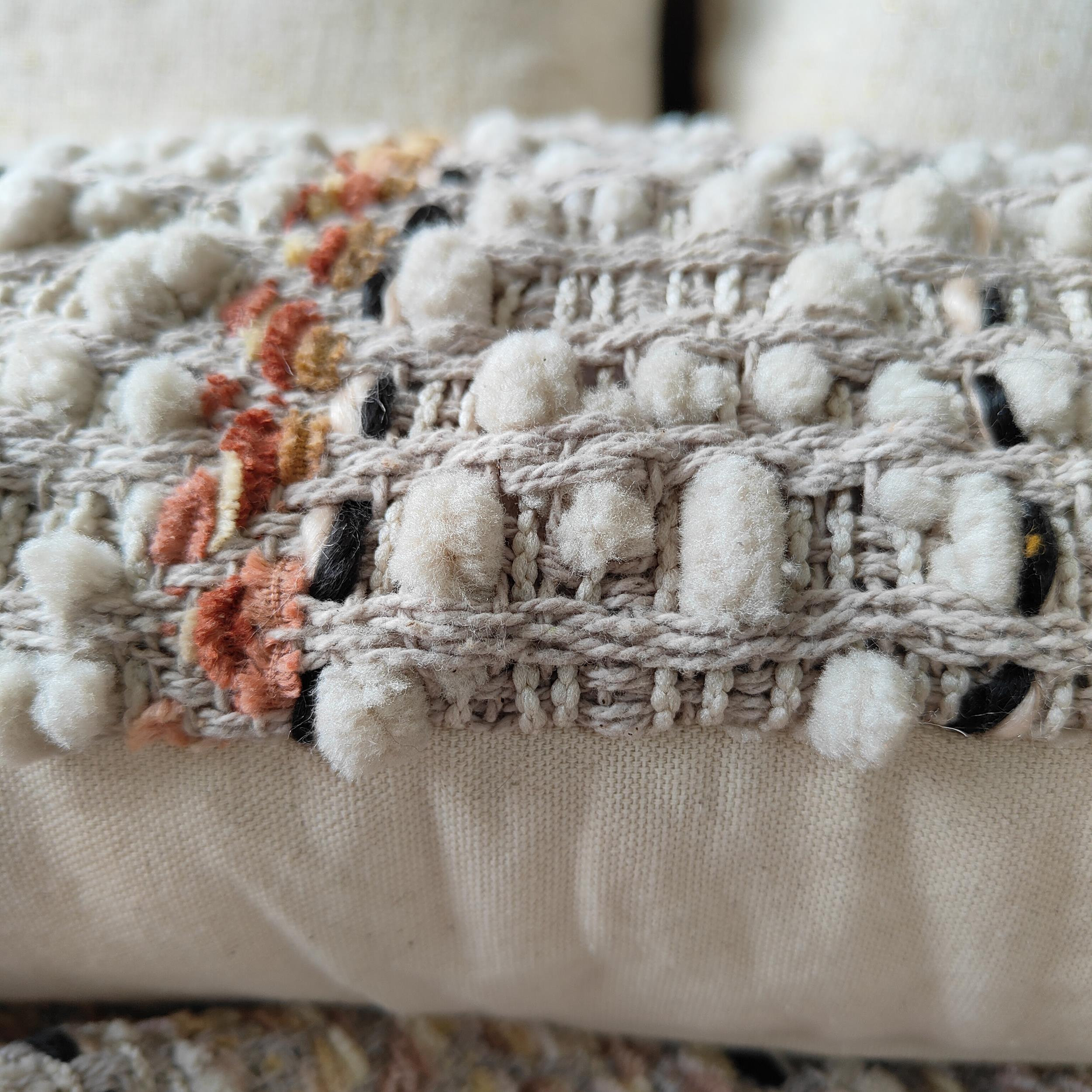 May include: Close-up of a beige throw pillow with a woven texture and white pom-poms. The pillow has a subtle orange and brown stripe pattern.