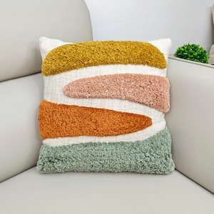 May include: Square decorative pillow with a white background and colorful, textured, abstract shapes. The shapes are in shades of gold, pink, orange, and green. The pillow is on a white surface.