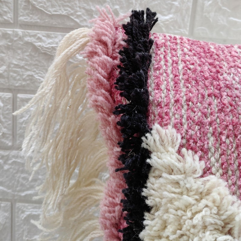May include: A pink, white, and black woven throw pillow with fringe detailing. The pillow has a textured, woven design and a soft, fluffy texture.