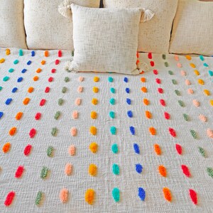 Puede incluir: Una manta blanca con un patrón de pompones coloridos. La manta tiene filas de pompones en tonos de azul, verde, naranja, rosa y amarillo.