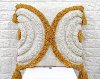 May include: Square decorative pillow with a textured off-white background and a mustard yellow geometric design. The pillow features a tufted pattern and is adorned with tassels at each corner, adding a bohemian flair to home decor.