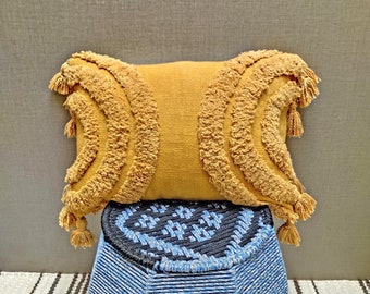 May include: A mustard yellow throw pillow with a fringe border and a woven design. The pillow is sitting on a blue and black woven stool.