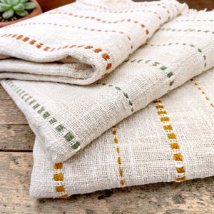 May include: A folded off-white woven textile featuring vertical stripes of embroidered stitching in shades of burnt orange, sage green, and mustard yellow. The fabric has a visible, coarse, and textured weave throughout the material.