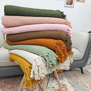 May include: A stack of waffle-weave blankets in various colors, including olive green, dusty rose, pink, brown, orange, sage green, cream, and mustard yellow. Each blanket has a fringed edge. The blankets are stacked on a wooden stool.