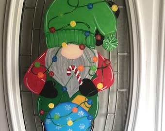 Puede incluir: Un gnomo de Navidad para colgar en la puerta con un sombrero verde, una camisa roja y una bola azul. El gnomo está decorado con luces de colores y un bastón de caramelo.