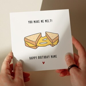 May include: A square white greeting card featuring a cartoon illustration of a grilled cheese sandwich with melted yellow cheese stretching between two halves. The text reads YOU MAKE ME MELT! and HAPPY BIRTHDAY NAME above a small red heart.