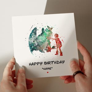 Op de afbeelding: Een witte wenskaart met een aquarel illustratie van een draak en een kind. De draak is voornamelijk groen en blauwgroen, terwijl het kind rood is. De kaart leest "HAPPY BIRTHDAY" en "*NAME*" met een klein rood hart.