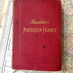 May include: A red hardcover book titled Baedeker's Northern France with gold embossed lettering on the front cover. The book features a textured fabric binding with subtle vertical and horizontal embossed lines and several small black spots.