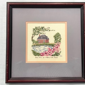 May include: A framed cross-stitch embroidery depicting the Rice Mill at Middleton Place. The scene features a brick building, a pond, trees, and pink flowers on a cream fabric background. The text RICE MILL at MIDDLETON PLACE is stitched below.