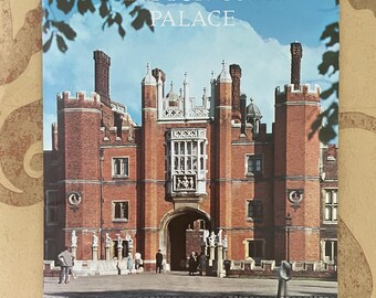 May include: A postcard featuring Hampton Court Palace. The image shows the palace's red brick facade, arched entrance, and multiple chimneys against a blue sky. The text "HAMPTON COURT PALACE" is printed at the top of the postcard.