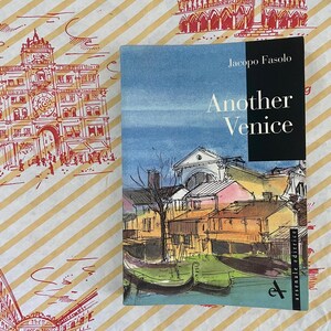 May include: A book titled "Another Venice" by Jacopo Fasolo. The cover features a watercolor illustration of Venetian buildings and canals. The book rests on a patterned surface with architectural sketches and text in French and Japanese.