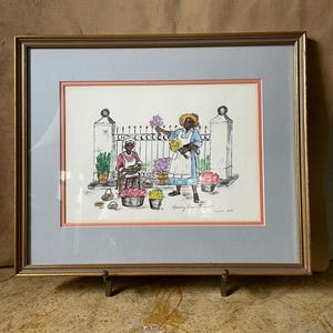 May include: Framed watercolor painting of two women selling flowers. The women are wearing hats and dresses. The painting is set in front of a white picket fence. The text "Rosemary Green Street" and "June 1985" is visible in the bottom right corner.