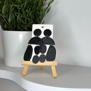 May include: Black abstract dangle earrings with gold flecks. The earrings are displayed on a small wooden easel. The earrings have a variety of shapes, including circles, half-circles, and oblong shapes. The earrings are attached to a white card.