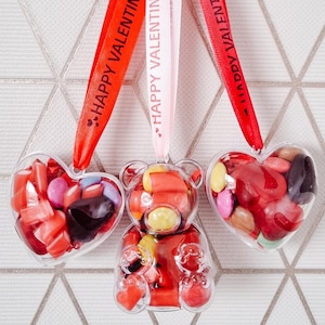 K&ouml;nnte beinhalten: Drei transparente Plastik-Valentinstags-Anh&auml;nger gef&uuml;llt mit bunten S&uuml;&szlig;igkeiten. Die Anh&auml;nger sind in Form eines Herzens, eines B&auml;ren und eines weiteren Herzens. Sie h&auml;ngen an roten und rosa B&auml;ndern mit dem Aufdruck "Happy Valentine's Day".