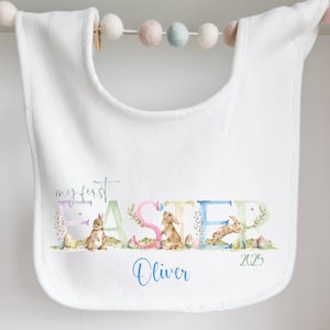 May include: White bib with the words "my first Easter" and the letters "EASTER" in pastel colors with bunnies and eggs. The name "Oliver" and the year "2025" are written below.