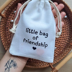 May include: A small white fabric drawstring pouch featuring the black printed text "little bag of friendship" with a small black heart icon below the text. The bag is made of a woven material and includes two off-white cotton drawstrings at the top.