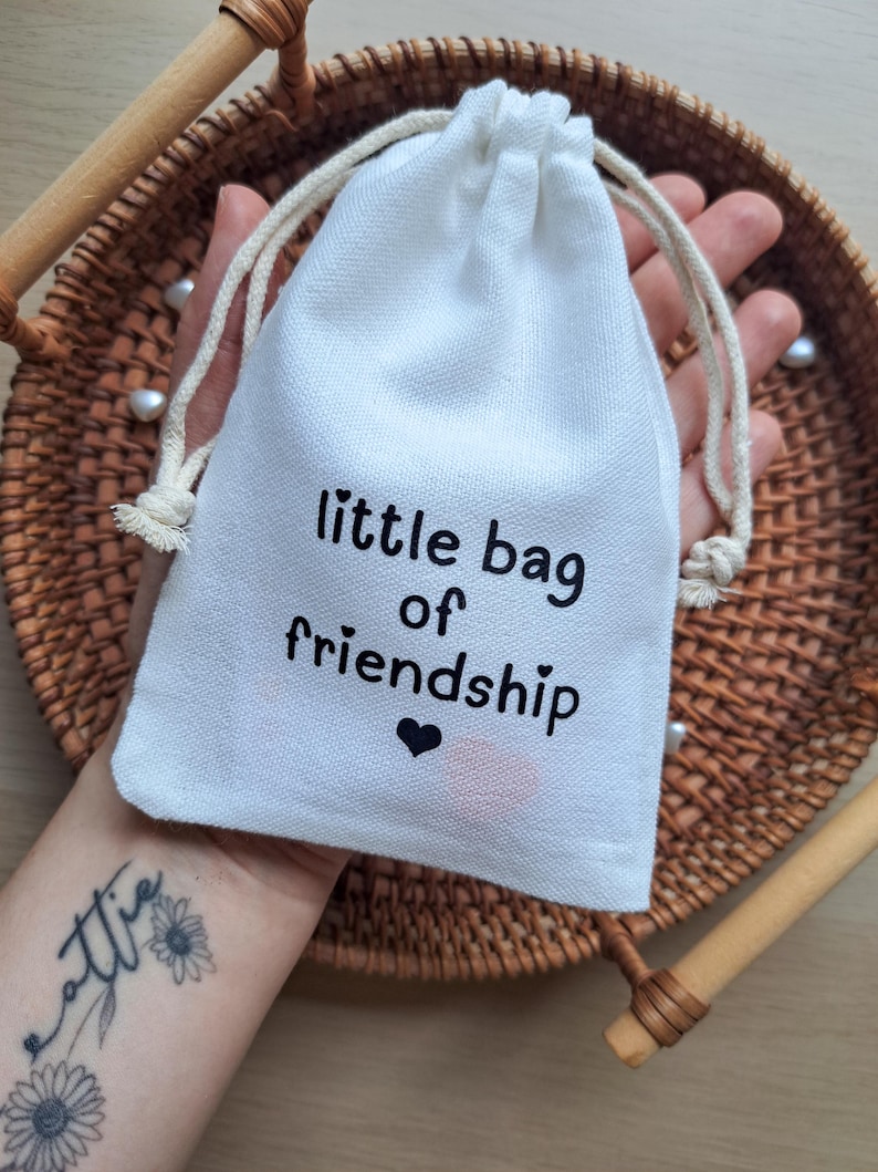 May include: A small white fabric drawstring pouch featuring the black printed text "little bag of friendship" with a small black heart icon below the text. The bag is made of a woven material and includes two off-white cotton drawstrings at the top.