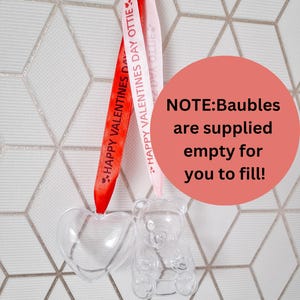 K&ouml;nnte beinhalten: Zwei transparente Plastik-Ornamente, eines herzf&ouml;rmig und das andere b&auml;renf&ouml;rmig, h&auml;ngen an roten und rosa B&auml;ndern mit dem aufgedruckten Text "HAPPY VALENTINES DAY OTTIE".  HINWEIS: Die Kugeln werden leer geliefert, damit Sie sie f&uuml;llen k&ouml;nnen!