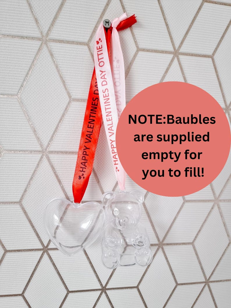 K&ouml;nnte beinhalten: Zwei transparente Plastik-Ornamente, eines herzf&ouml;rmig und das andere b&auml;renf&ouml;rmig, h&auml;ngen an roten und rosa B&auml;ndern mit dem aufgedruckten Text "HAPPY VALENTINES DAY OTTIE".  HINWEIS: Die Kugeln werden leer geliefert, damit Sie sie f&uuml;llen k&ouml;nnen!