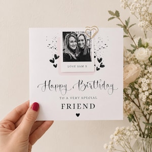 Könnte beinhalten: Eine quadratische, weiße Geburtstagskarte mit einem schwarz-weißen Foto von zwei Personen, das mit einer goldenen herzförmigen Büroklammer befestigt ist. Die Karte enthält den Text Happy Birthday to a very special FRIEND und schwarze Herzen.