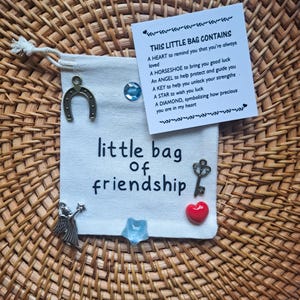 May include: A small, off-white drawstring bag with the words "little bag of friendship" printed on it. The bag is decorated with a horseshoe charm, a key charm, a heart, a star, a diamond, and an angel. A card lists the contents and their meanings.