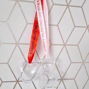 K&ouml;nnte beinhalten: Zwei transparente Plastik-Anh&auml;nger, einer in Herzform und der andere in B&auml;renform, h&auml;ngen an rosa und roten B&auml;ndern mit dem Text "Happy Valentines Day Ottie" darauf.