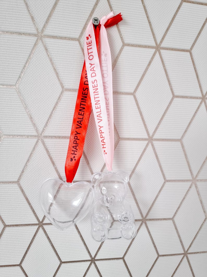 K&ouml;nnte beinhalten: Zwei transparente Plastik-Anh&auml;nger, einer in Herzform und der andere in B&auml;renform, h&auml;ngen an rosa und roten B&auml;ndern mit dem Text "Happy Valentines Day Ottie" darauf.