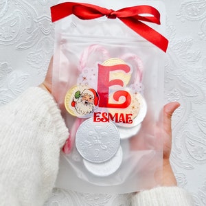 May include: A clear plastic bag with a red ribbon tied around the top. The bag contains candy canes, chocolate coins, and a sticker with the letter "E" and the name "Esmae".