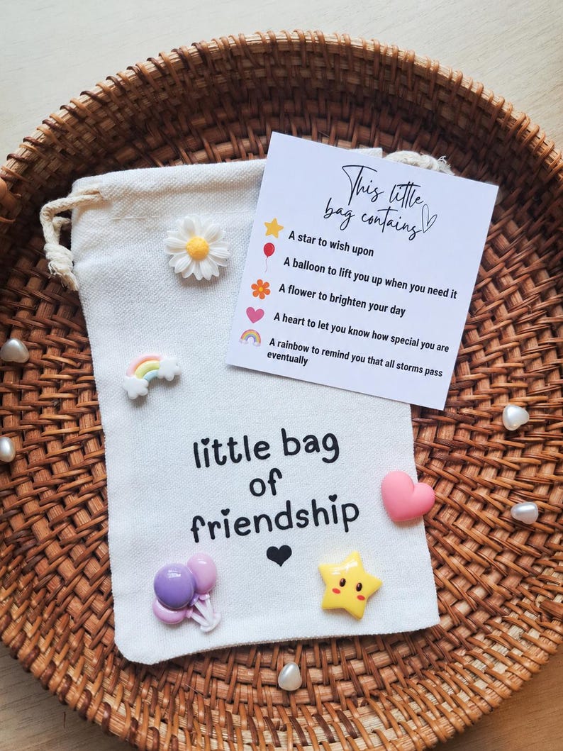 May include: A cream-colored drawstring pouch with the text 'little bag of friendship' and a black heart icon. It is decorated with small resin charms including a daisy, rainbow, balloons, heart, and star, and includes a card titled 'This little bag contains'.