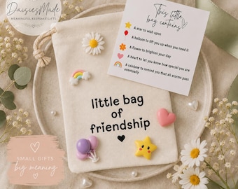May include: A cream-colored fabric drawstring pouch with the text "little bag of friendship" printed in black. The pouch is decorated with small resin charms including a daisy, a rainbow, two balloons, and a yellow star, plus a card.