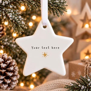 Puede incluir: Un adorno de cerámica blanca en forma de estrella que cuelga de una cinta blanca. La superficie frontal presenta el texto impreso en negro "Your text here" sobre un pequeño icono de estrella dorada con líneas radiantes.